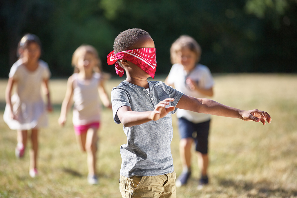 Ein Junge hat die Augen mit einem roten Kopftuch verbunden. Er hat die Arme tastend vorgestreckt. Im Hintergrund laufen drei Kinder auf den Jungen zu.