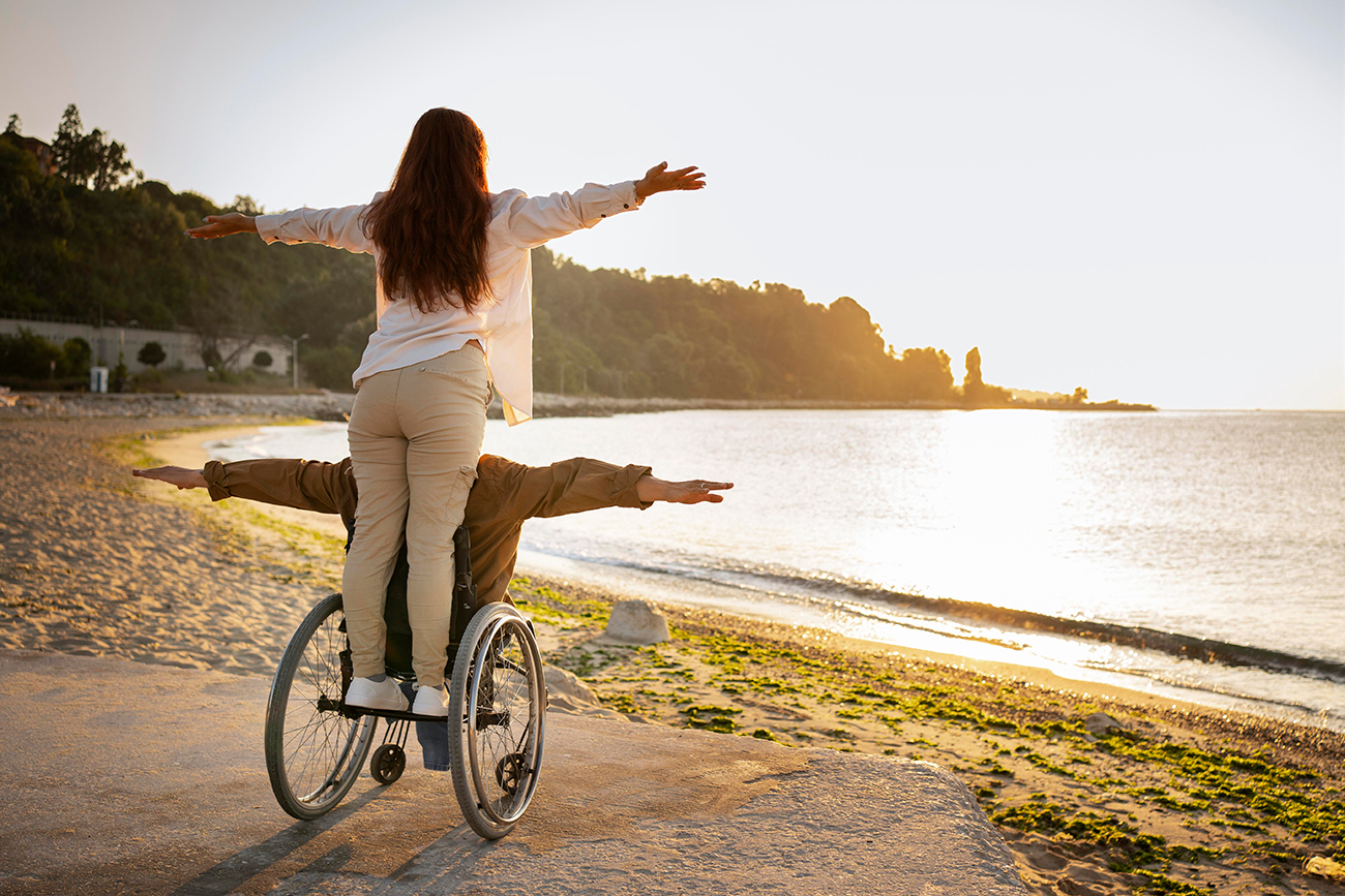 Website barrierefrei machen! So weit klar. Aber warum und wie? 1 Ein Mann und eine Frau befinden sich voller Freude an einem Ufer und Blicken auf einen kleinen See. Die Sonne glitzert auf der Oberfläche des Wassers. Der Mann sitzt im Rollstuhl, die Frau steht hinten auf einem Tritt auf dem Rollstuhl. Beide breiten die Arme aus, als könnten sie fliegen.